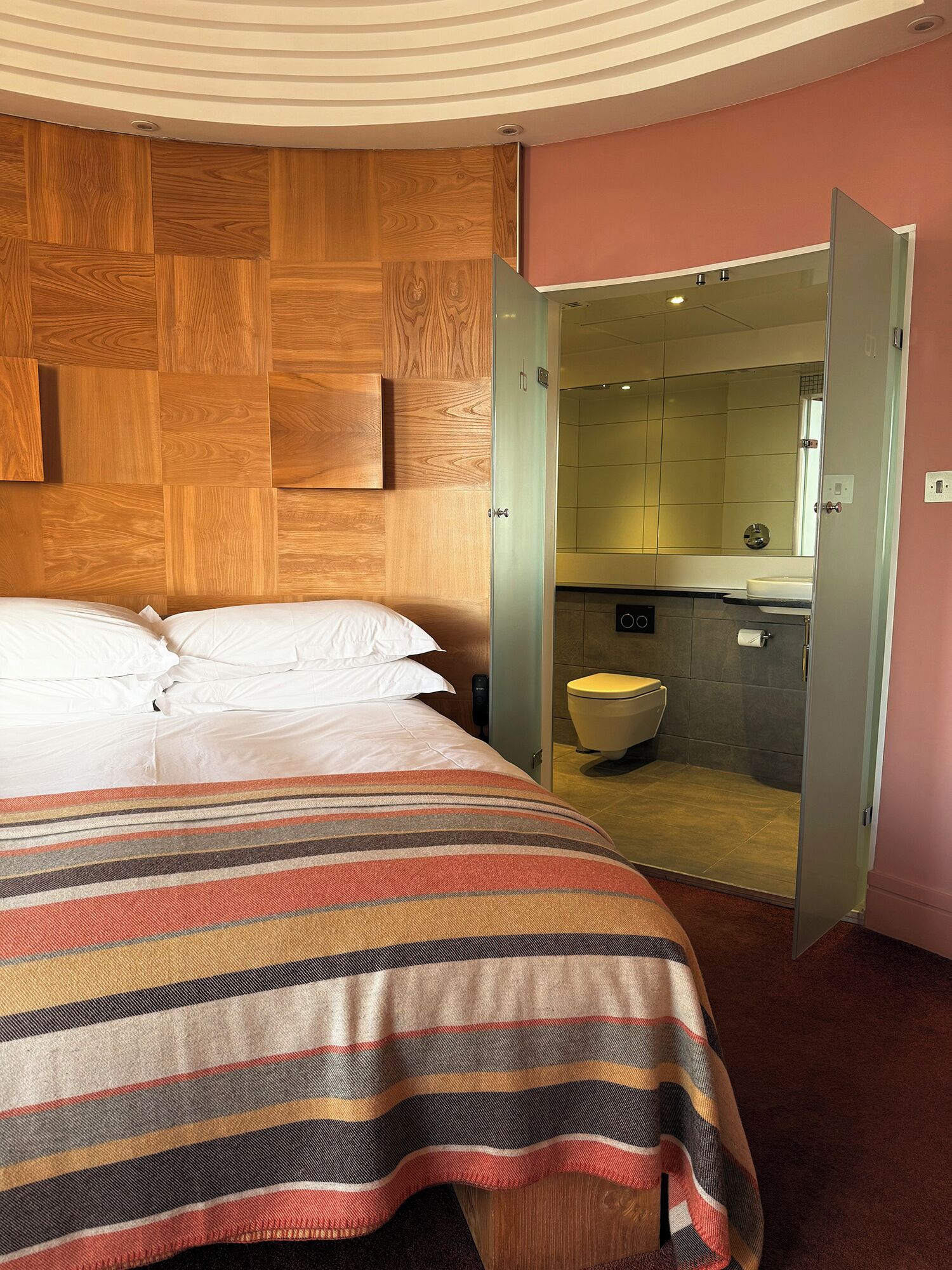 A hotel room with a bed, a striped blanket, a wooden headboard, and an open door leading to a bathroom with a toilet and a mirror.