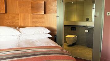 A hotel room with a bed, a striped blanket, a wooden headboard, and an open door leading to a bathroom with a toilet and a mirror.