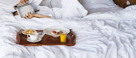 Una persona recostada en la cama con una bandeja de desayuno que incluye croissants, una taza de café y jugo de naranja.