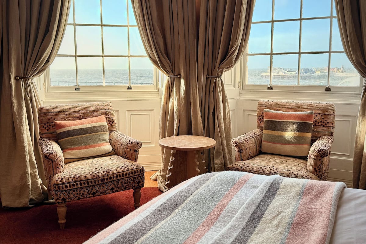 A room with two patterned armchairs, a small round table, and a bed with a striped blanket.