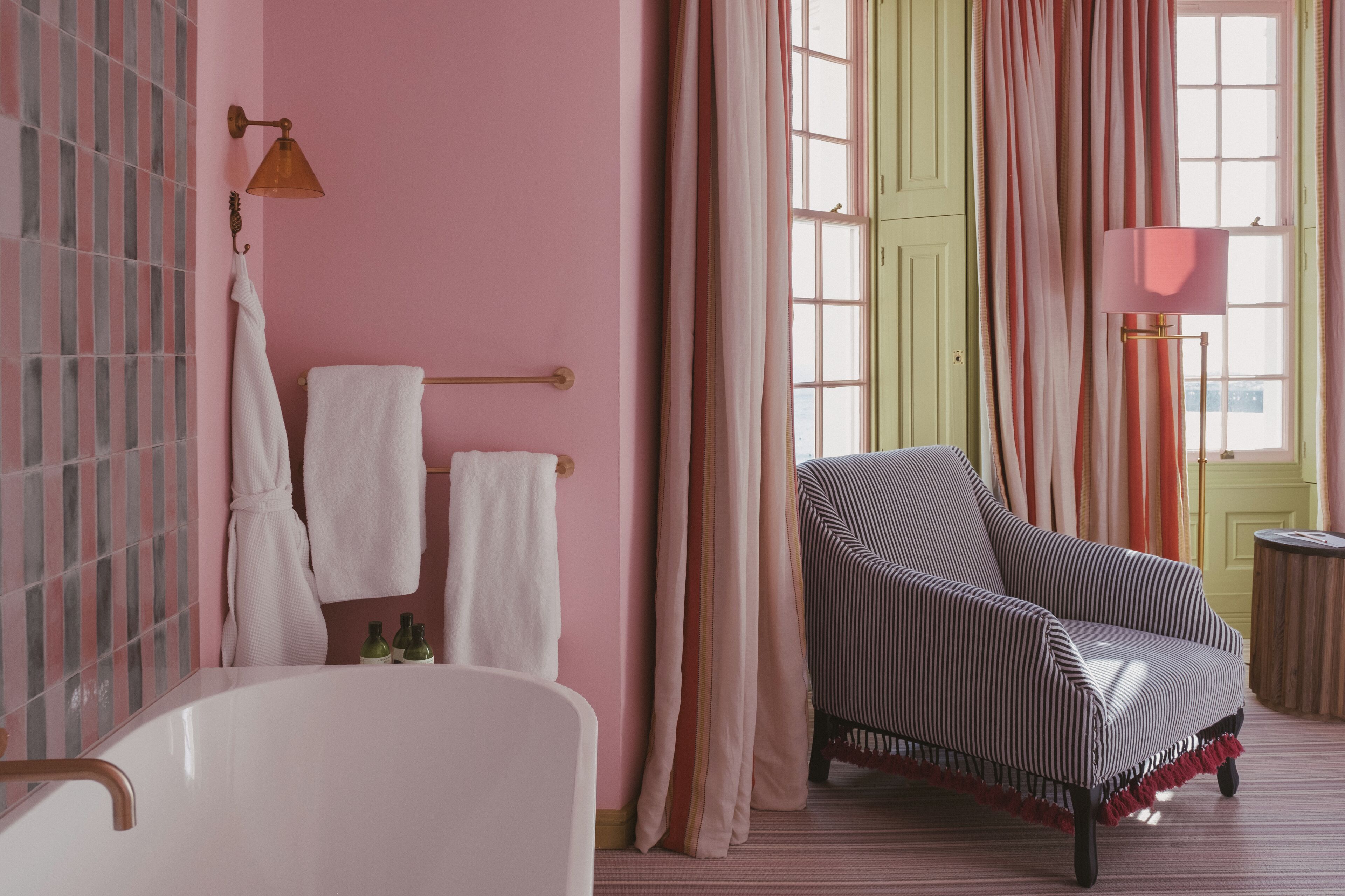 A bathroom with a white bathtub, striped armchair, and pink walls.