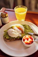 Un plato de desayuno con tostada de aguacate, un huevo escalfado y un tazón pequeño de fruta.
