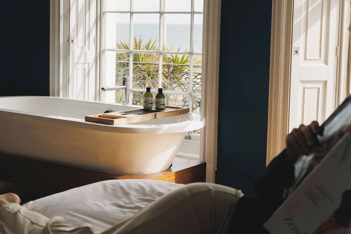 A bathroom with a freestanding bathtub, a window with a view of the sea, and a wooden tray on the edge of the tub.