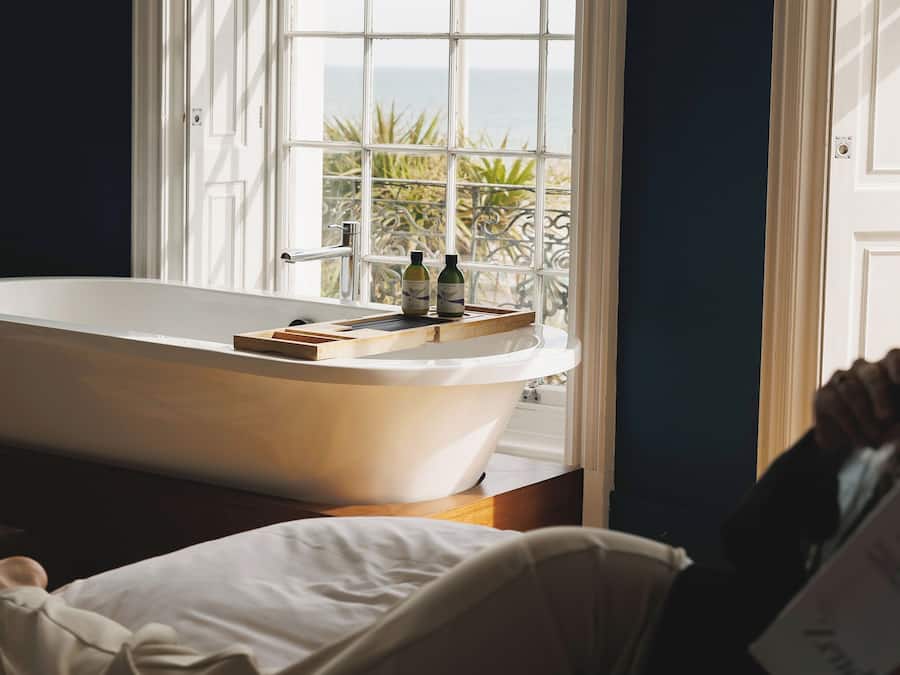 A bathroom with a freestanding bathtub, a window with a view of the sea, and a wooden tray on the edge of the tub.