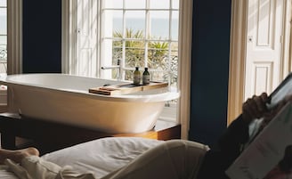 A bathroom with a freestanding bathtub, a window with a view of the sea, and a wooden tray on the edge of the tub.