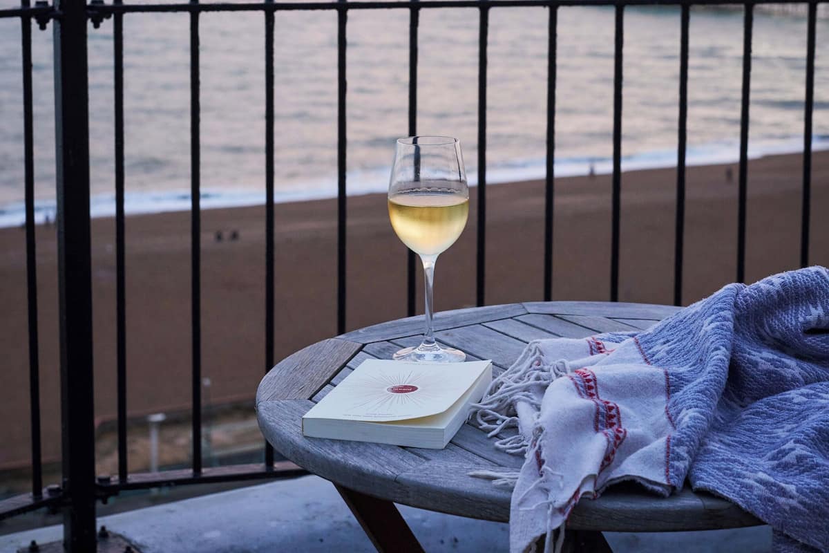 A table with a glass of white wine, a book, and a blanket on a balcony overlooking the sea.