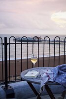 A table with a glass of white wine, a book, and a blanket on a balcony overlooking the sea.