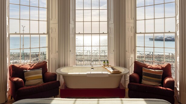 A bedroom with a large window, a bathtub, two armchairs, and a bed. - Drakes of Brighton (Brighton)