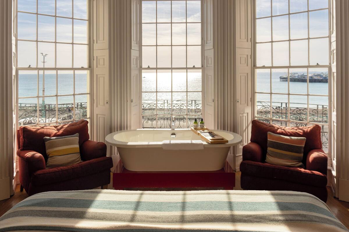 A bedroom with a large window, a bathtub, two armchairs, and a bed.