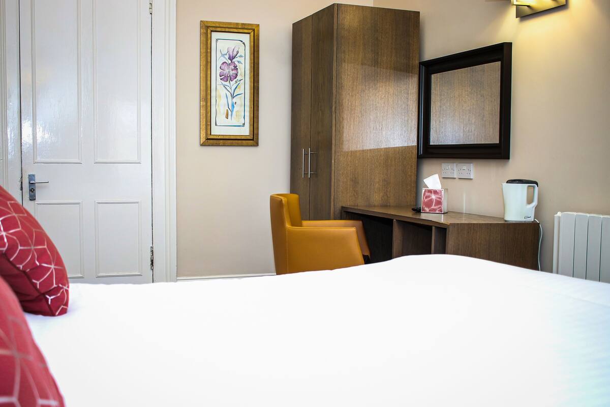 A hotel room with a bed, a yellow chair, a wooden wardrobe, a framed artwork, and a small table with a tissue box and a kettle.