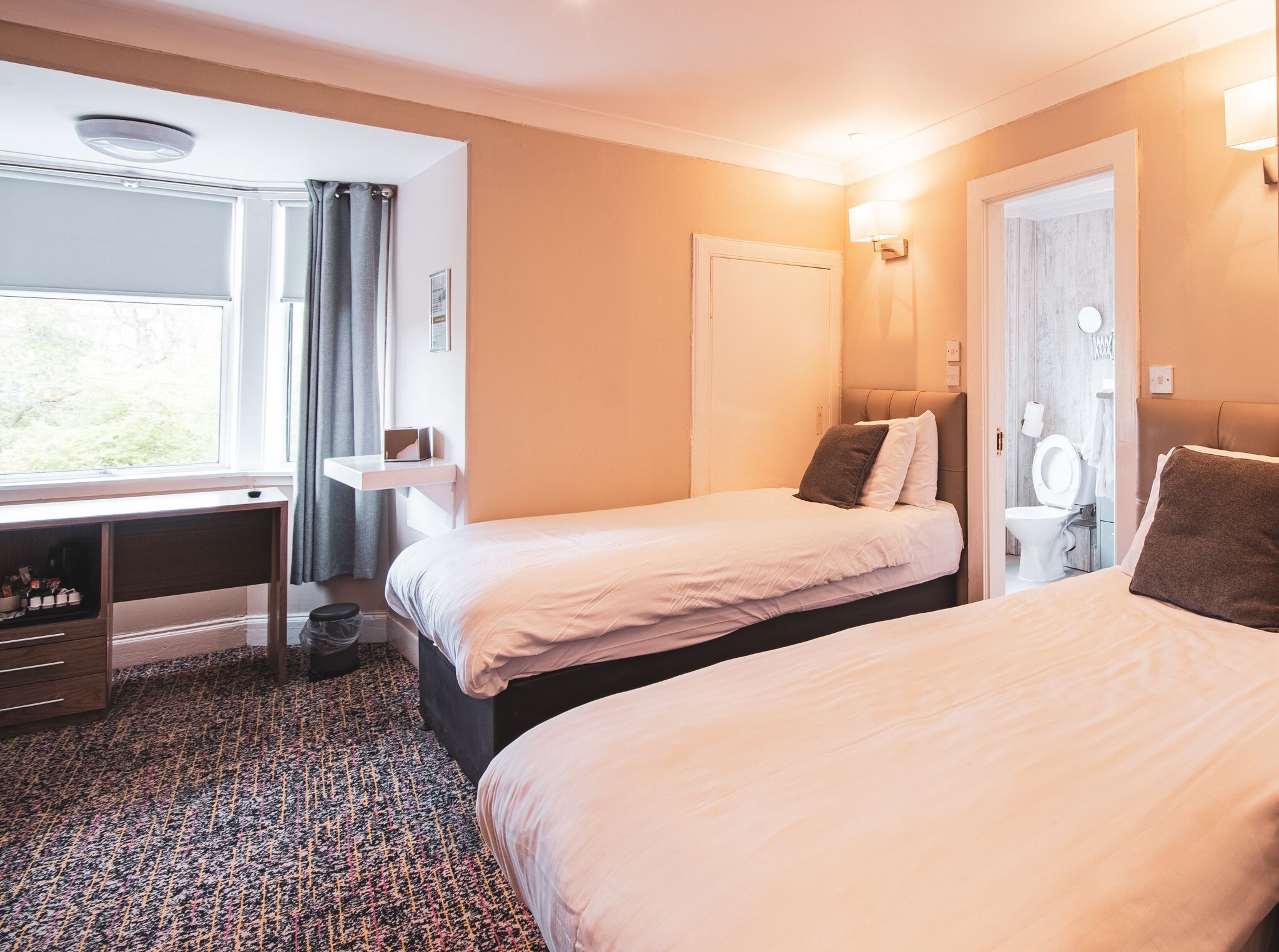 A hotel room with two beds, a desk, a window with curtains, a bathroom with a toilet and sink, and a patterned carpet.