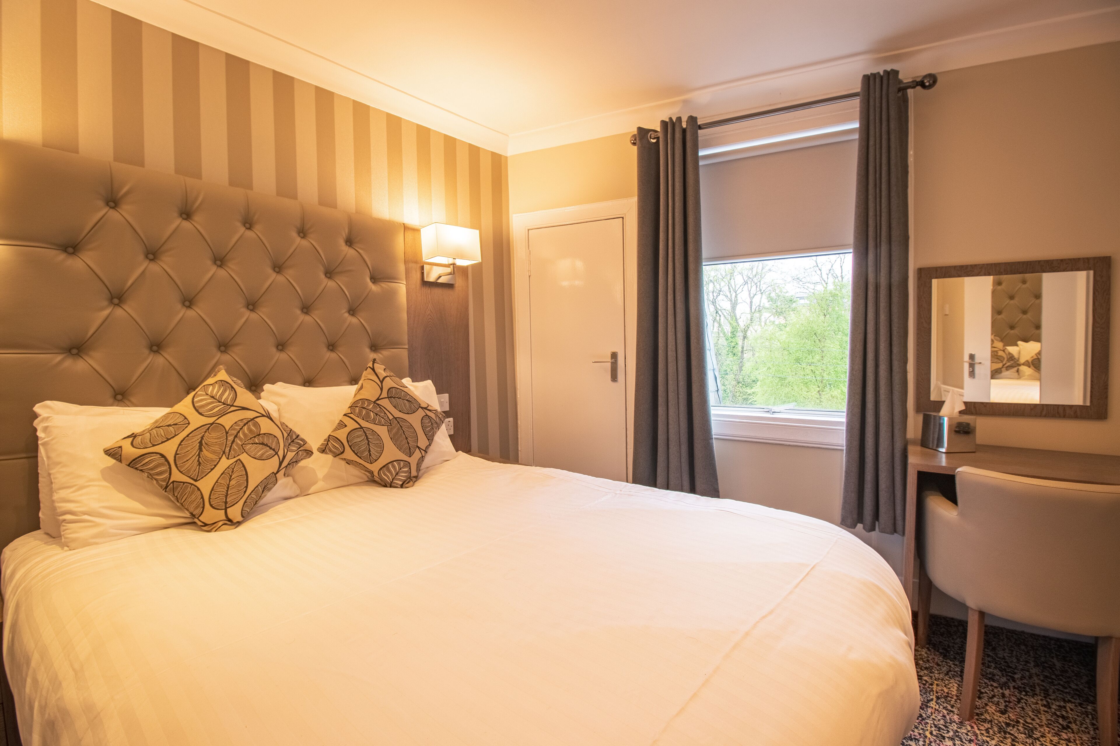 A hotel room with a tufted headboard, a bed with white linens and patterned pillows, a desk with a chair, and a window with curtains.