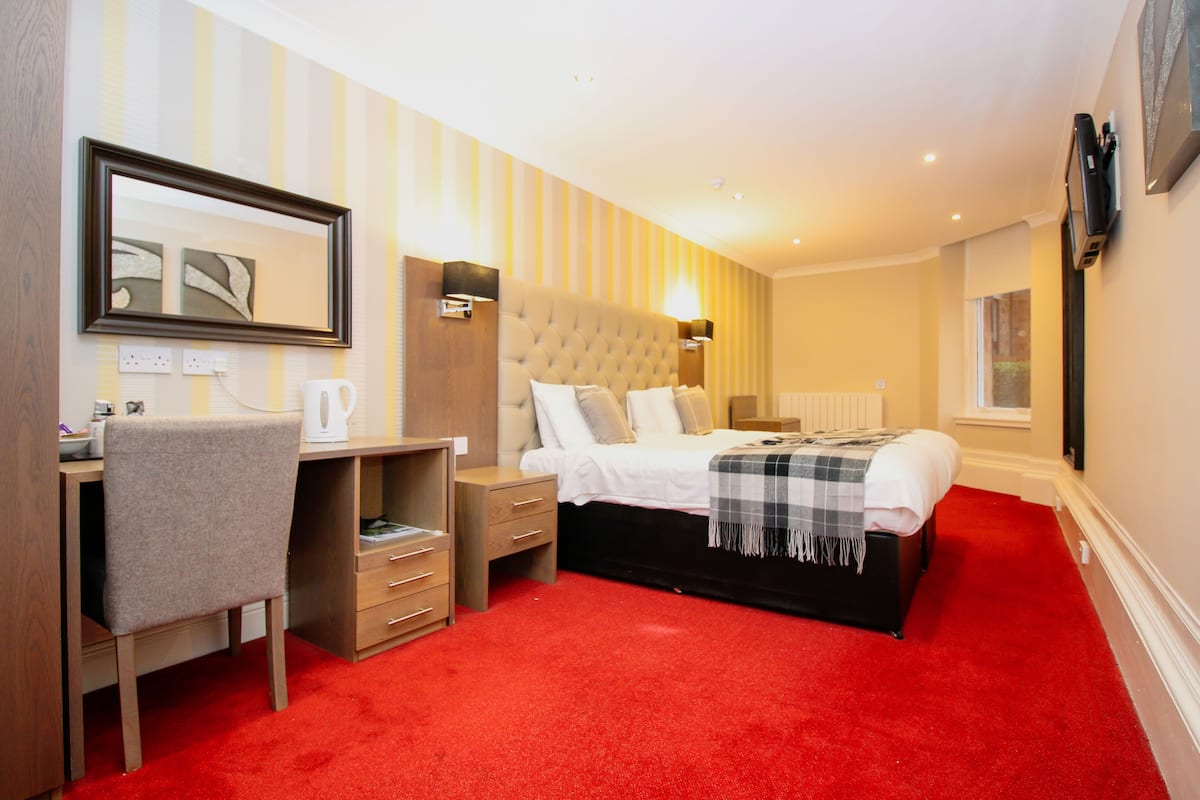 A hotel room with a bed, a desk, a chair, a nightstand, a lamp, and a mirror.