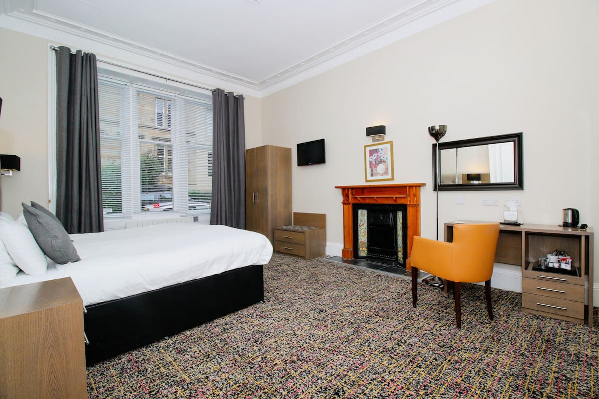 A hotel room with a bed, a desk, a chair, a fireplace, and a large window with curtains.