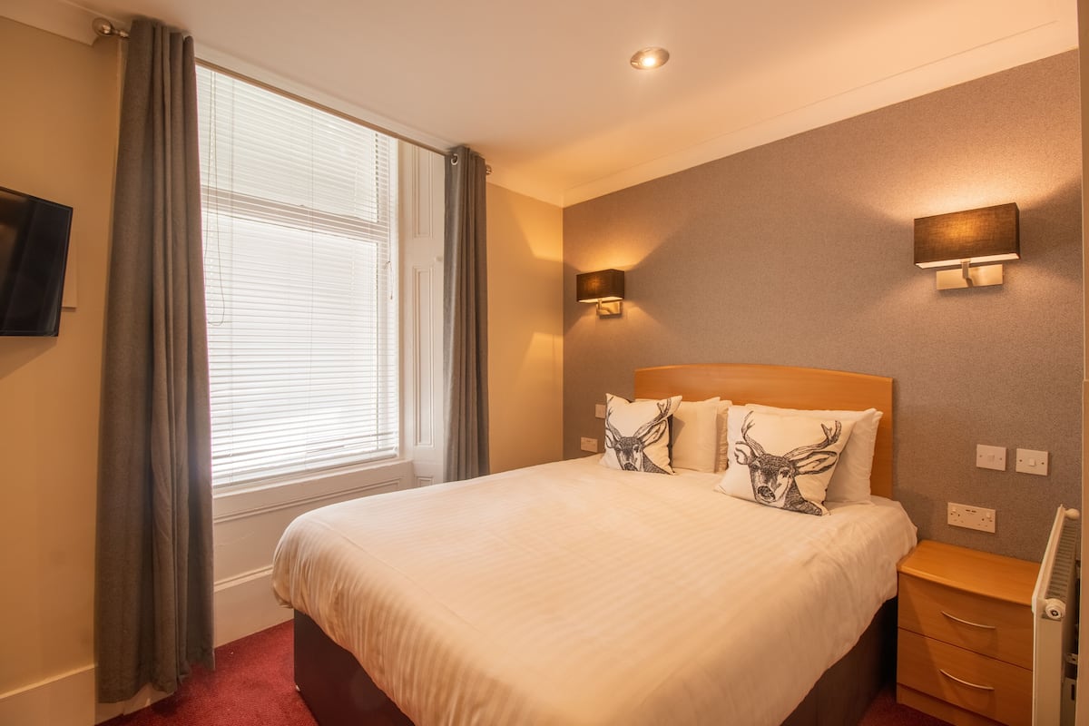 A hotel room with a large bed, two wall-mounted lamps, a flat-screen TV, and a window with curtains.