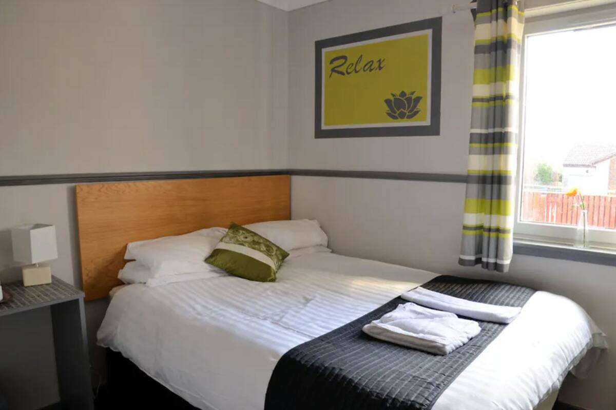 A neatly made bed with white linens and a green pillow, a bedside table with a lamp, and a framed 'Relax' sign on the wall.