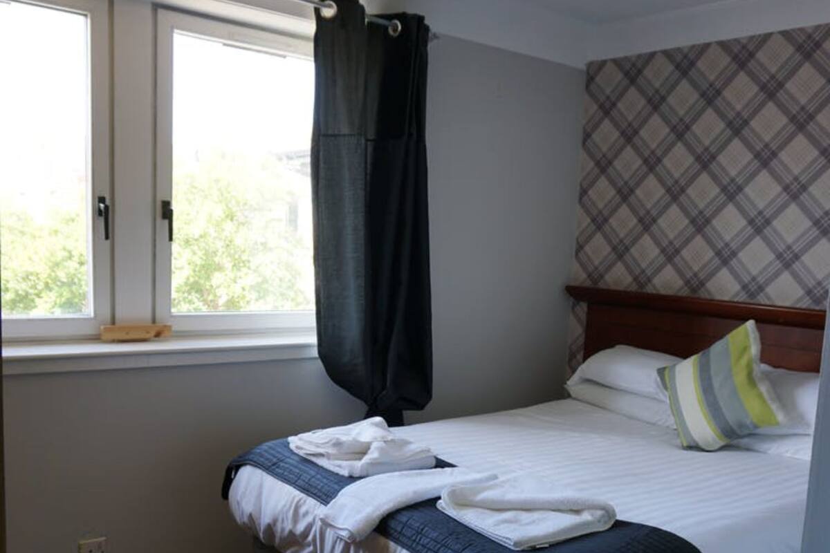 A neatly made bed with white and blue bedding, a plaid-patterned headboard, and a window with a black curtain.