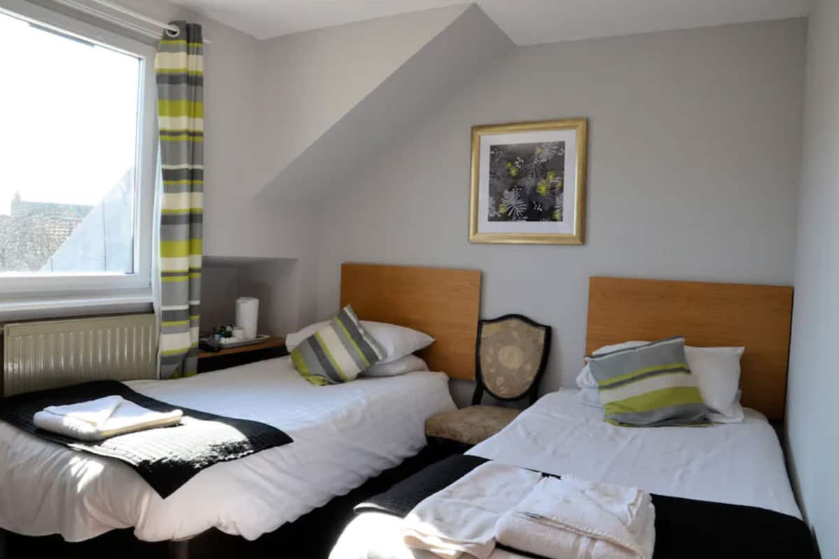 A hotel room with two beds, a window with curtains, a radiator, a framed picture on the wall, and a bedside table with a lamp.