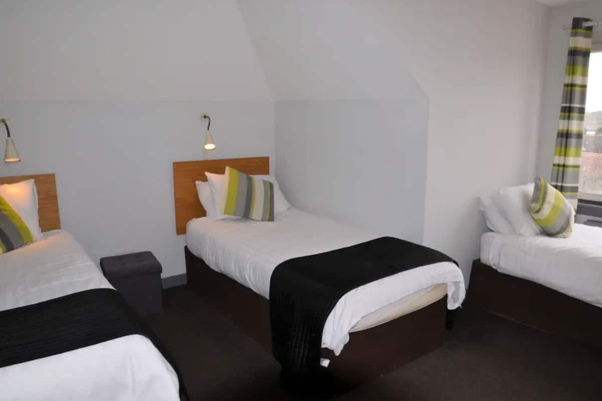 A hotel room with three single beds, each with a wooden headboard and white bedding. There are two bedside tables, one on each bed, and a small black trash bin. The room has white walls and a window with a striped curtain.