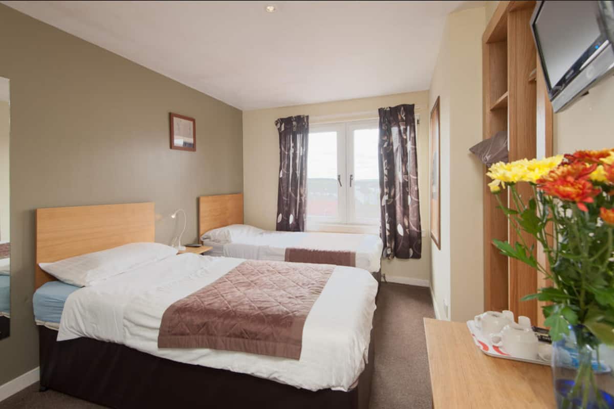 A hotel room with two beds, a TV, a desk with a vase of flowers, and a window with curtains.