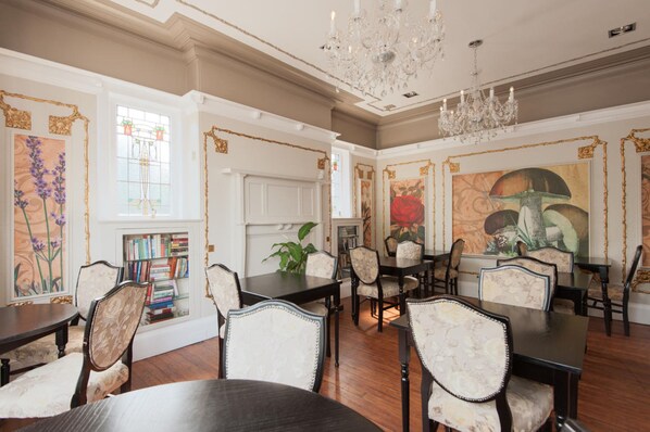 A dining room with a chandelier, wooden tables, and chairs. There is a bookshelf with books and a potted plant.