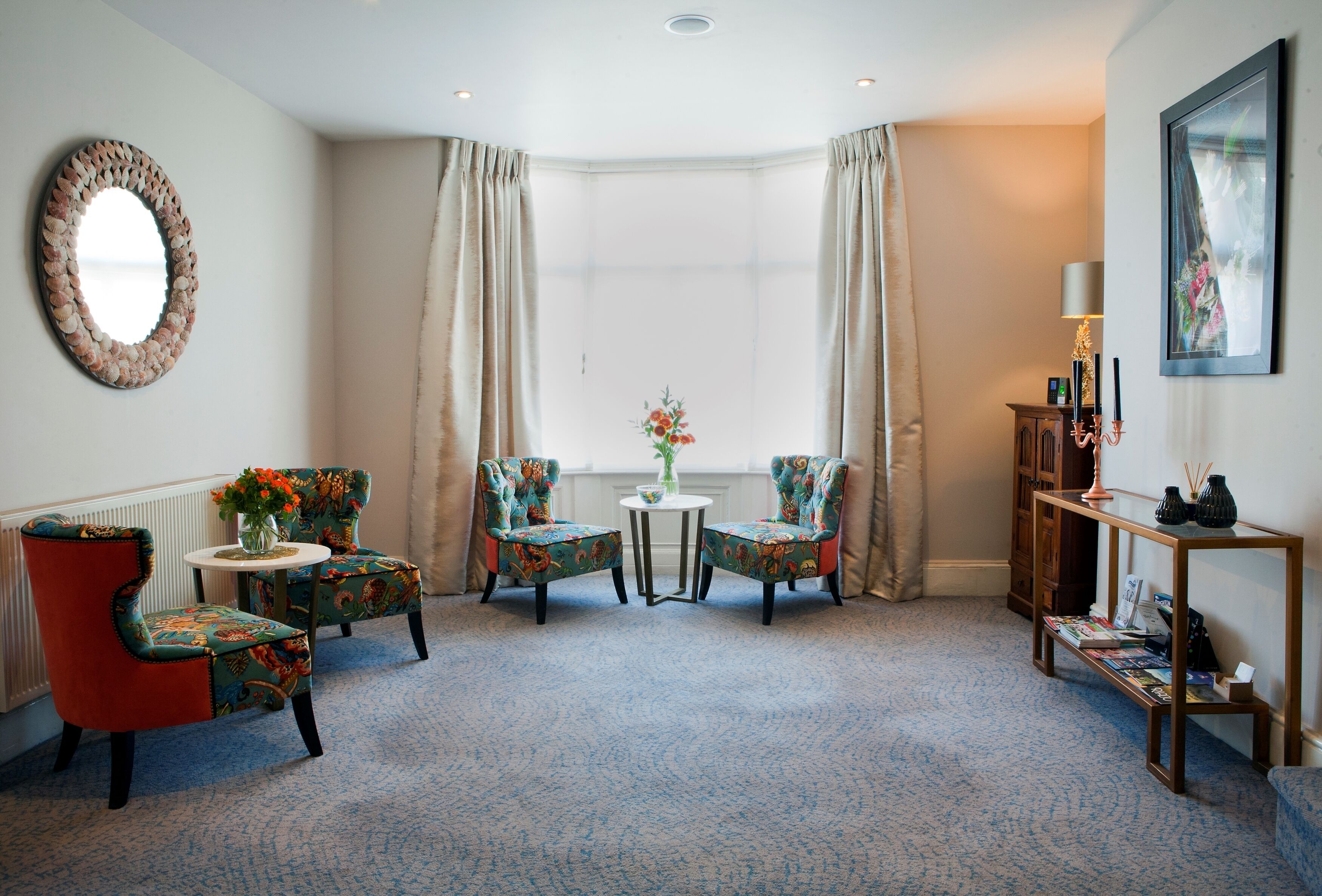 A room with two patterned armchairs, a small table with a vase of flowers, a wooden console table with books, and a large mirror on the wall.