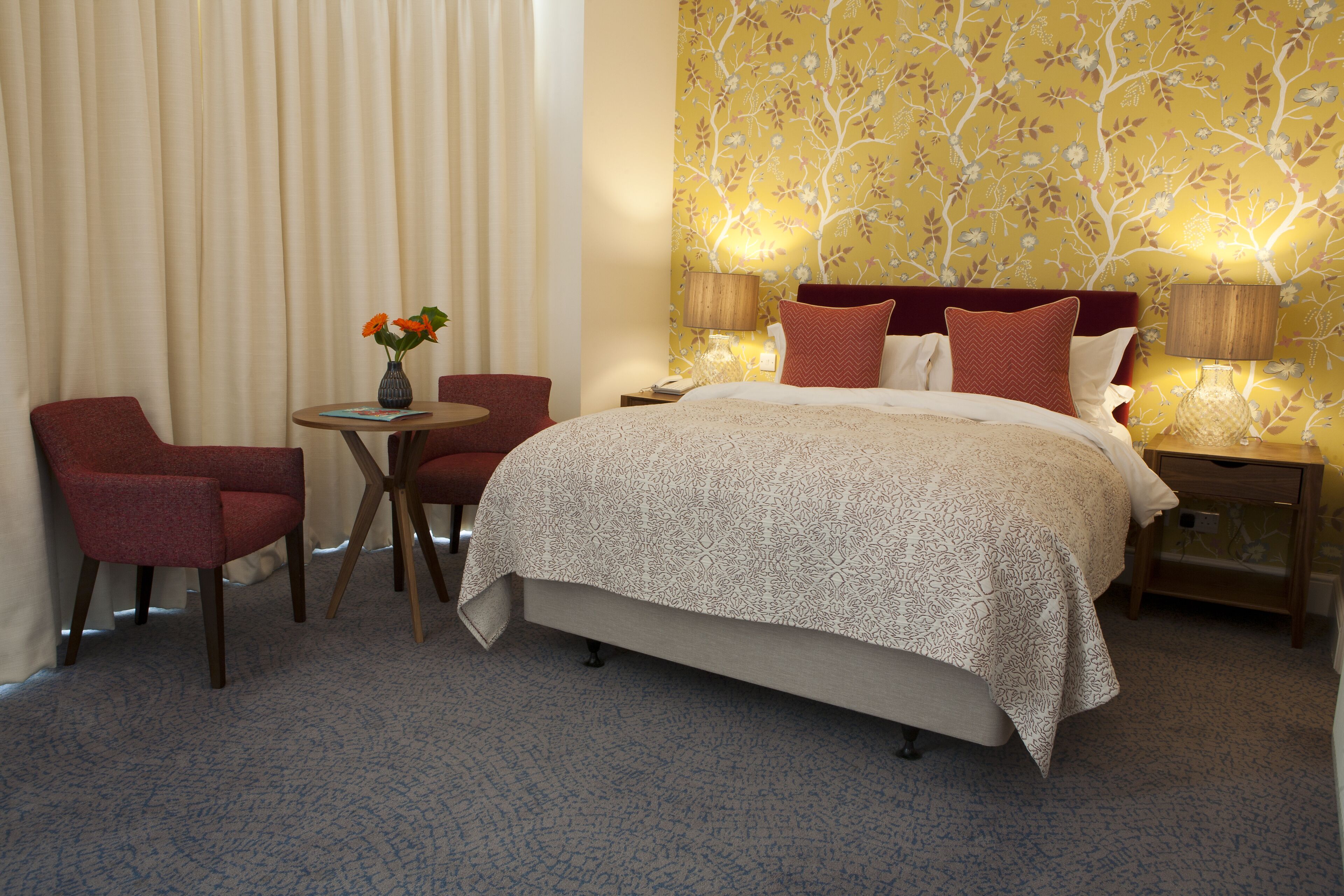 Habitación de hotel con cama, dos sillas, una mesita con un florero y una mesita de noche con lámparas.