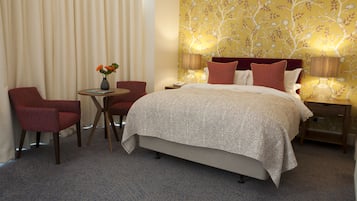 Habitación de hotel con cama, dos sillas, una mesita con un florero y una mesita de noche con lámparas.
