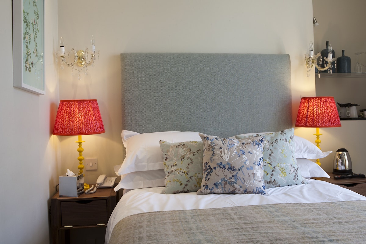 A hotel room with a bed, two bedside lamps, a nightstand with a phone, a small table with a kettle, and a wall with a framed picture.