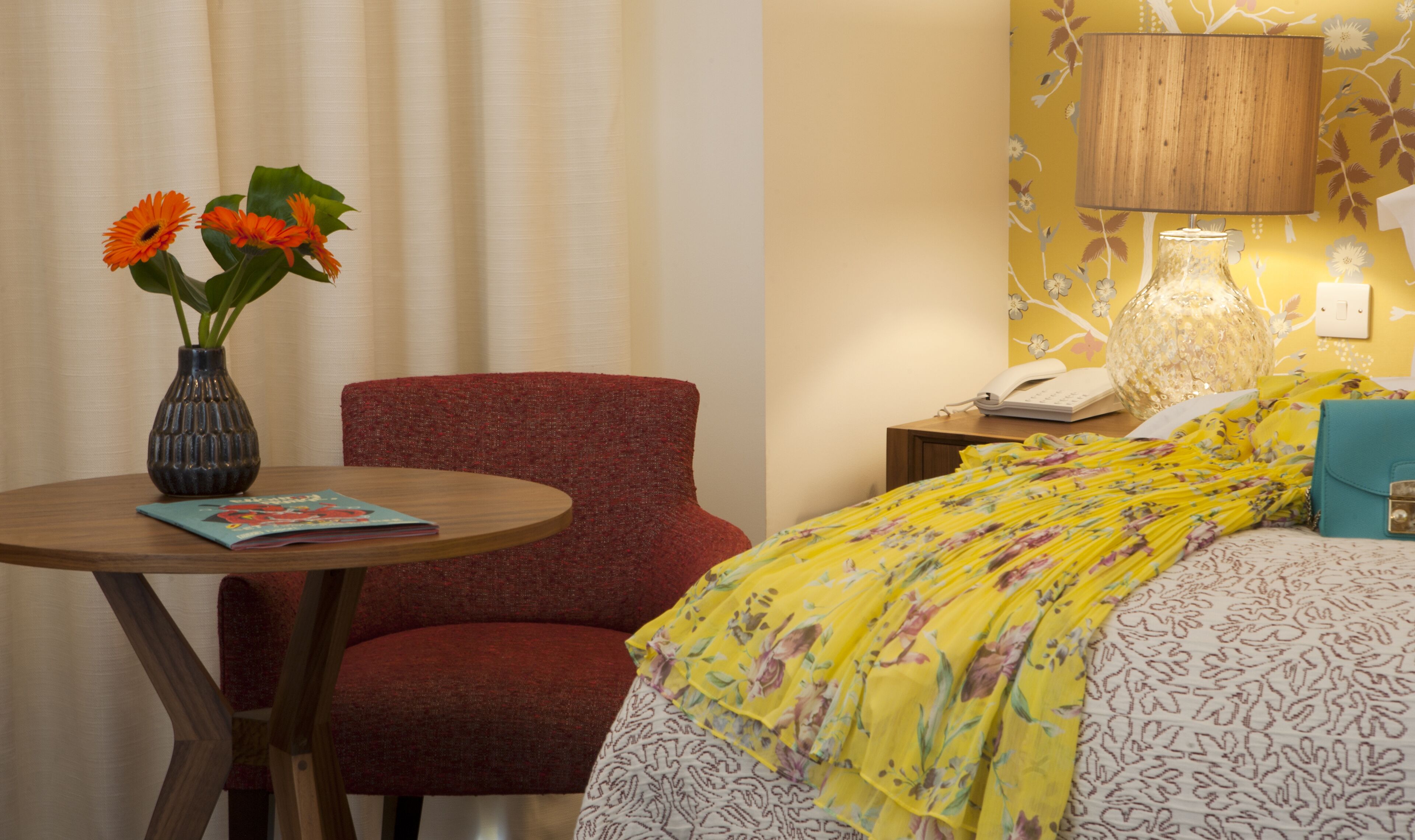 Habitación de hotel con un sillón rojo, una mesa redonda de madera con un jarrón de flores naranjas, una cama con colcha amarilla estampada, una lámpara y un teléfono en una mesita de noche.