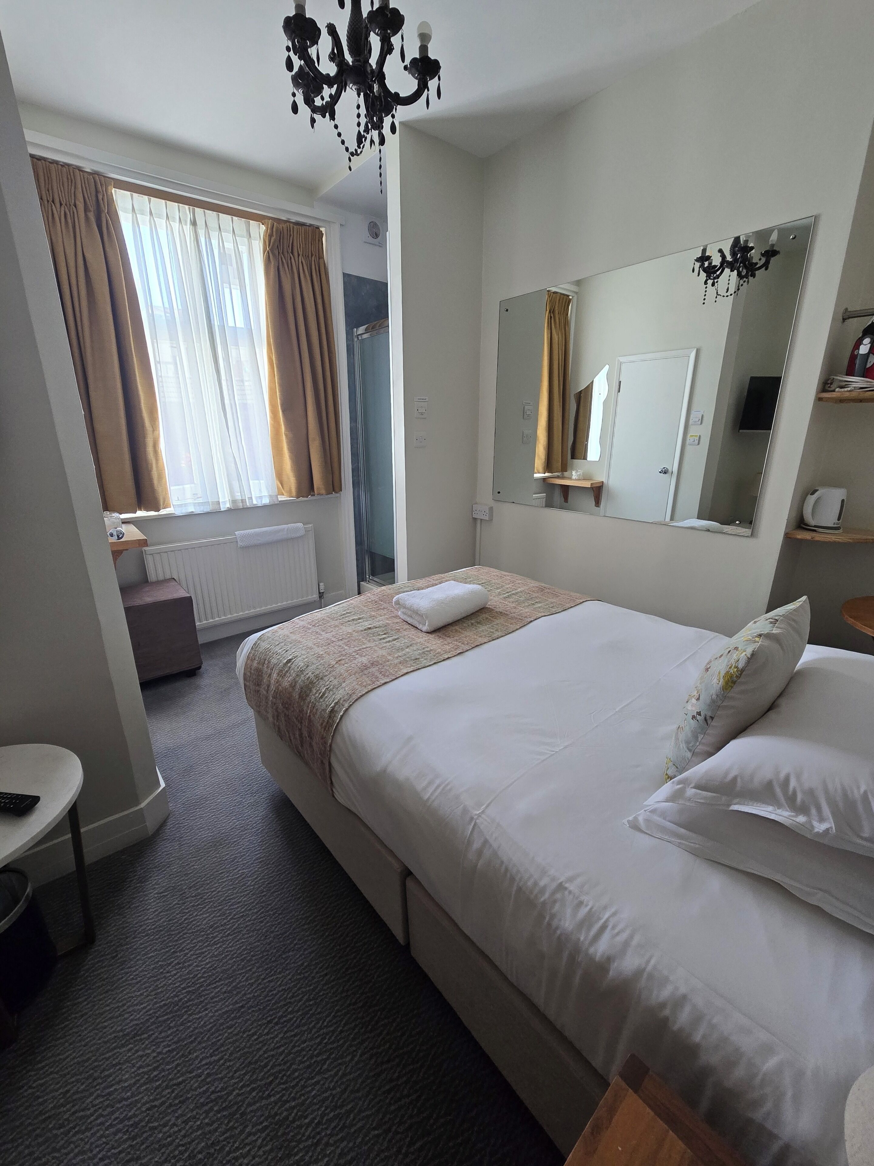 Habitación de hotel con una cama grande, una mesita de noche, un espejo, un ventanal con cortinas y una araña.