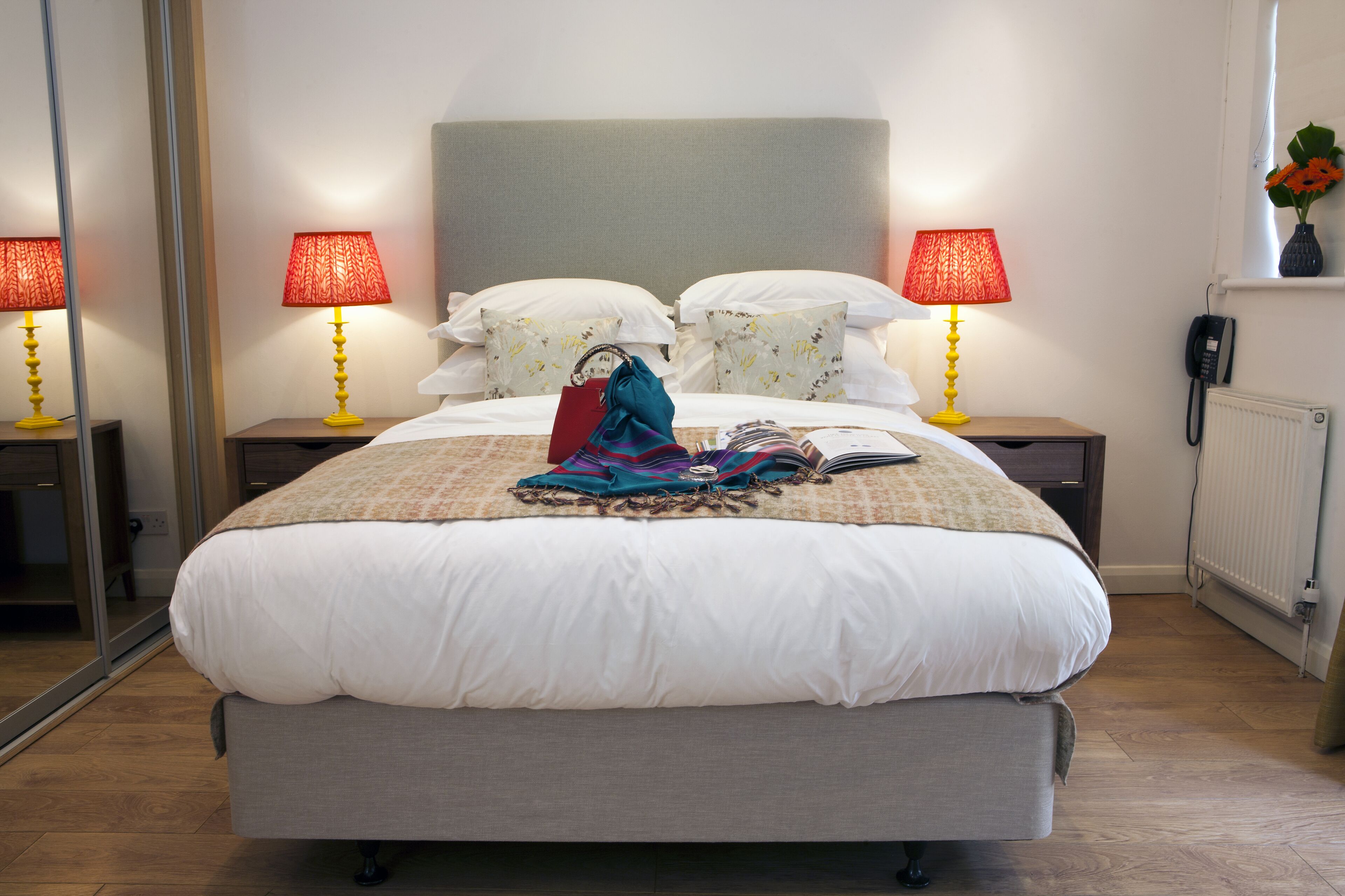 Cama bem arrumada com lençóis brancos, duas luminárias de cabeceira com abajures vermelhos, telefone ao lado da cama e espelho na parede à esquerda.