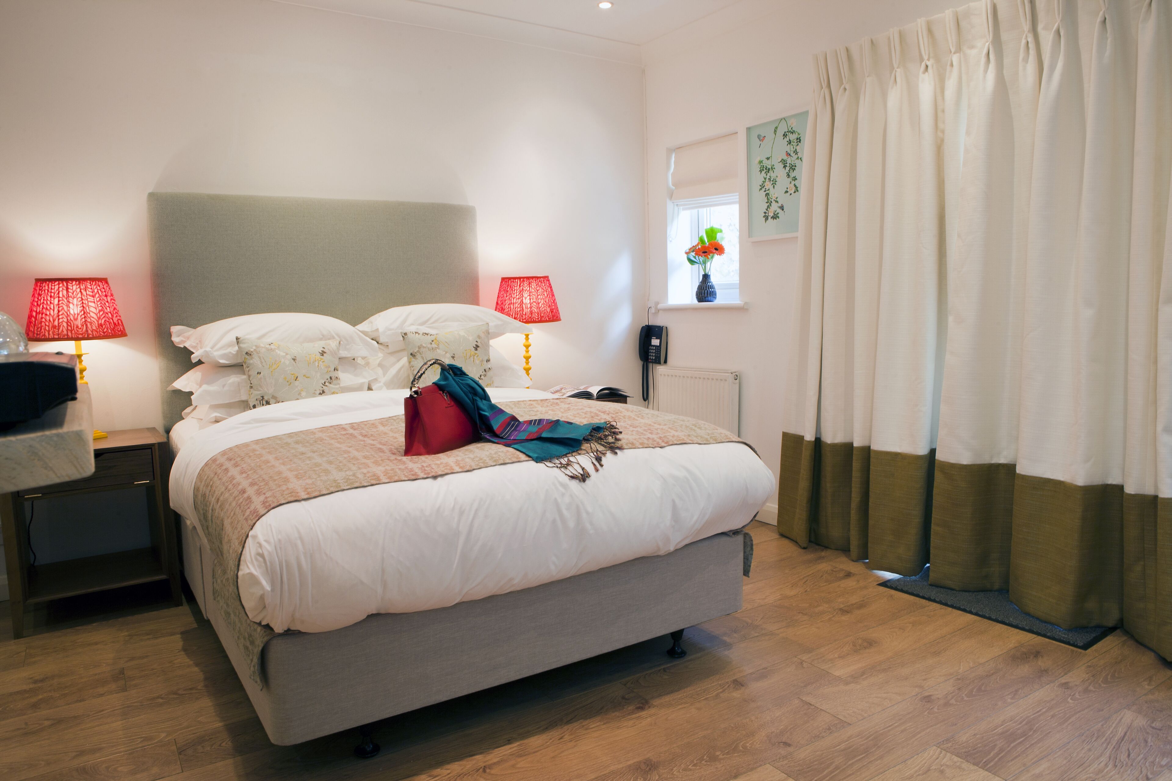 A neatly made bed with a headboard, two bedside tables with lamps, a window with a view, and a curtain.