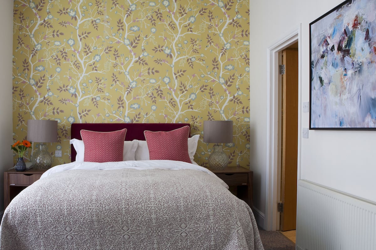 A hotel room with a floral wallpaper, a bed with a patterned bedspread, two bedside tables with lamps, and a painting on the wall.