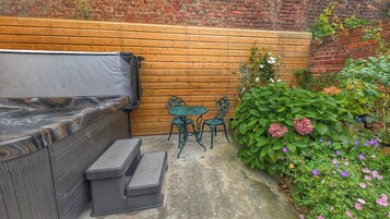 A hot tub with a wooden bench and steps, a small round table with chairs, and a brick wall with greenery and flowers.