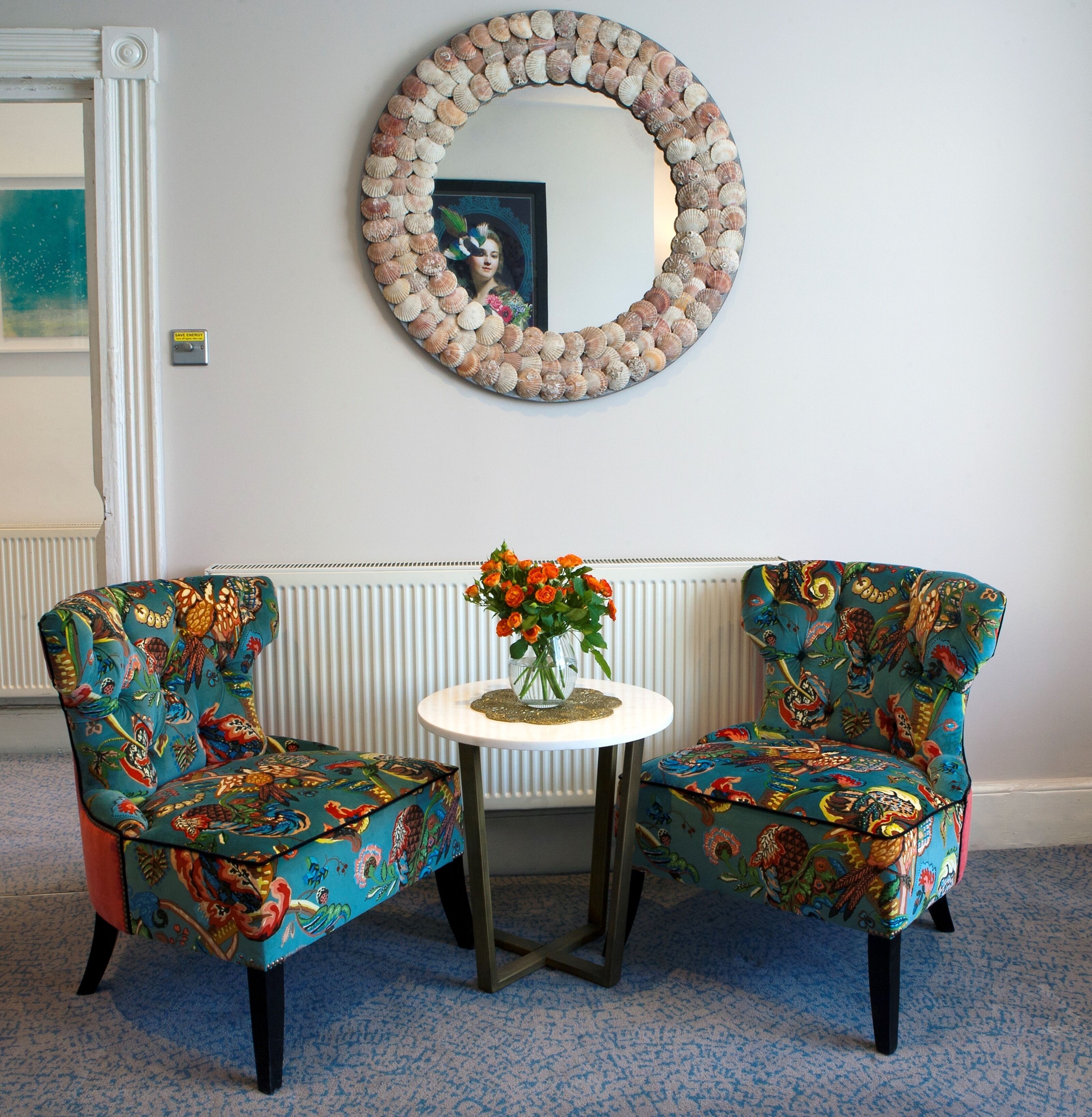 Una habitación con dos sillones estampados, una mesita redonda con un florero y un espejo decorativo en la pared.
