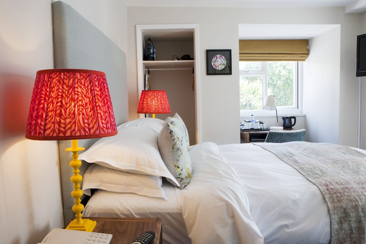 A hotel room with a bed, two bedside lamps, a bedside table with a phone and remote control, a window with a view, and a desk with a kettle.