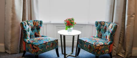 Two upholstered chairs with floral upholstery, a small round table with a vase of flowers, and a window with curtains.