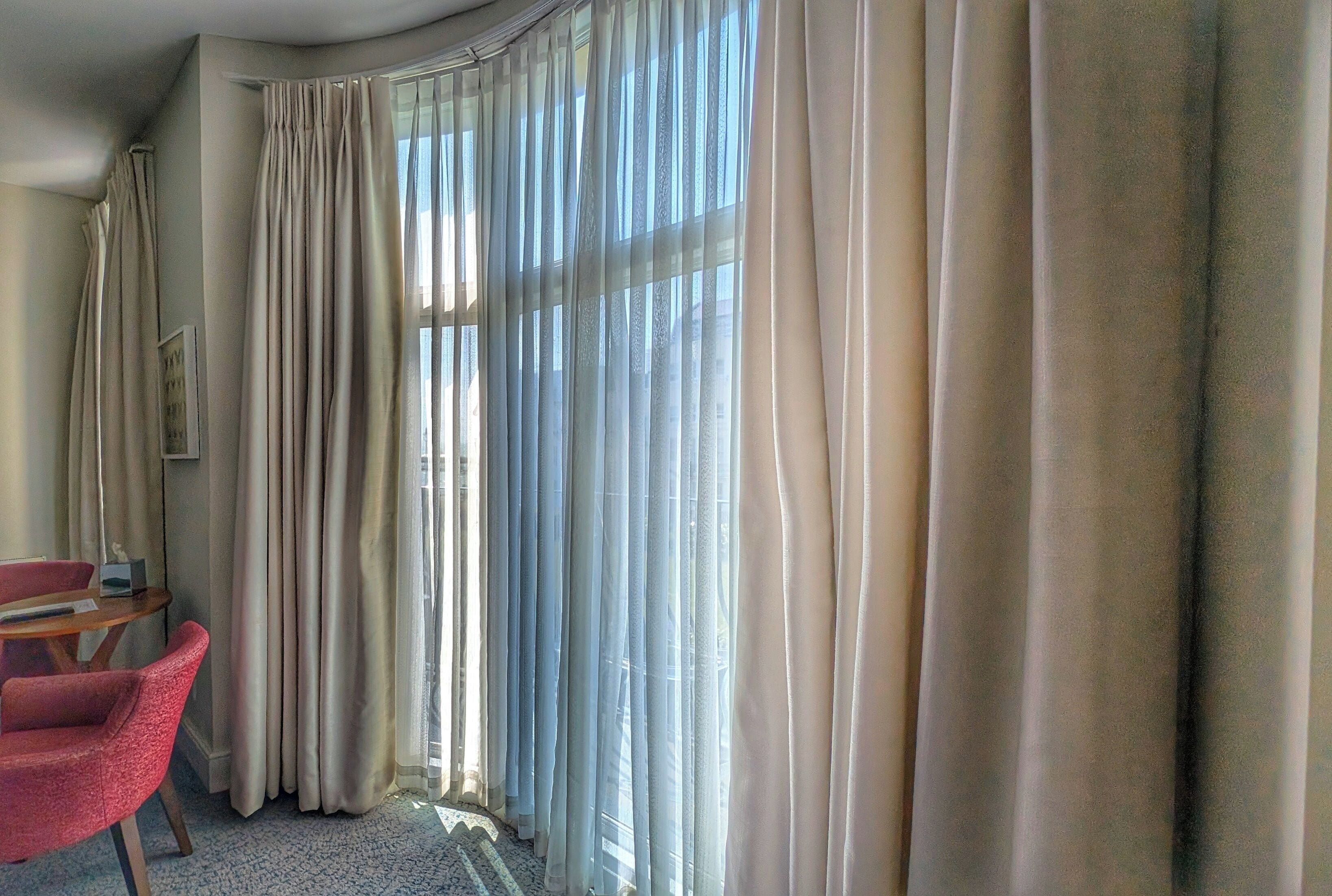 A hotel room with a large window covered by sheer curtains, a red chair, and a small table.