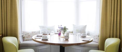 Habitación de esquina con una mesa redonda, dos sillas, un banco y una ventana con cortinas.