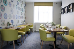 A dining area with yellow chairs and tables, a patterned wall, and a window with blinds. - Sea Spray (Brighton)