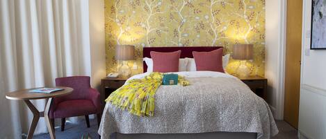 Habitación de hotel ordenada con una cama grande, dos lámparas de noche, un sillón rojo, una mesita redonda y un papel tapiz con motivos florales.