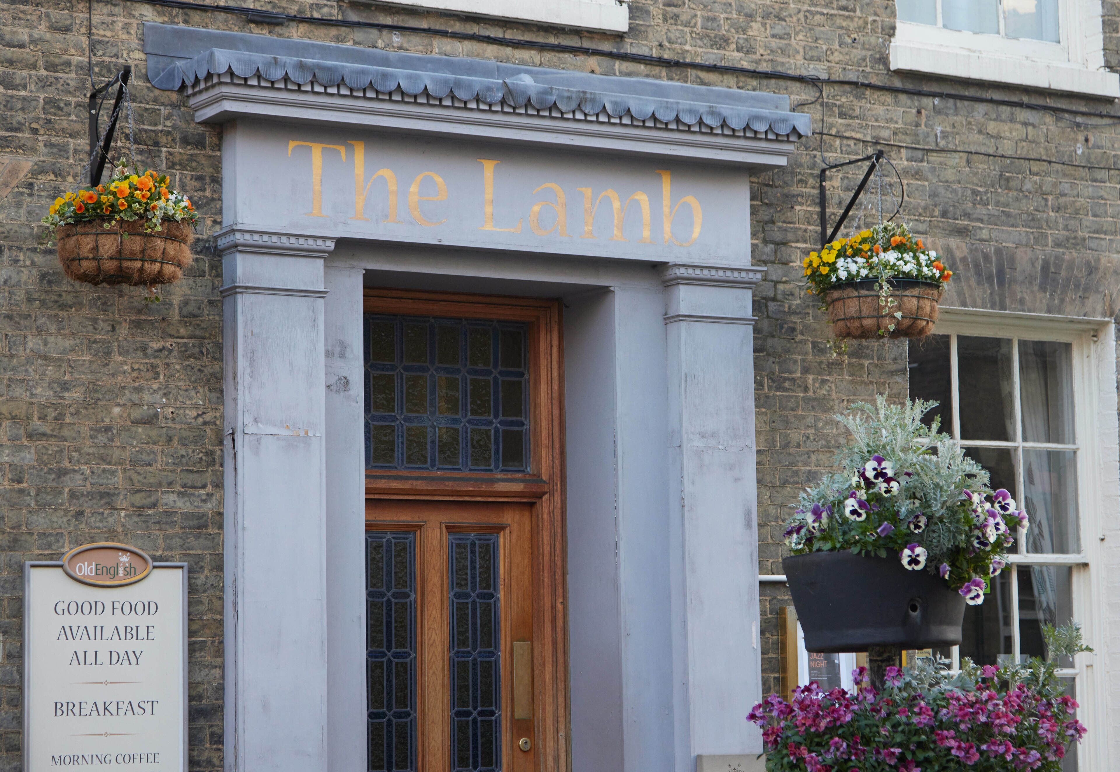 Un bâtiment sur lequel on peut lire l’inscription « The Lamb », et en dessous, une autre inscription indiquant : « NOURRITURE DE QUALITÉ SERVIE TOUTE LA JOURNÉE, PETIT-DÉJEUNER, CAFÉ DU MATIN ».