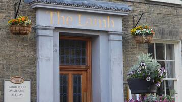 Un bâtiment sur lequel on peut lire l’inscription « The Lamb », et en dessous, une autre inscription indiquant : « NOURRITURE DE QUALITÉ SERVIE TOUTE LA JOURNÉE, PETIT-DÉJEUNER, CAFÉ DU MATIN ».