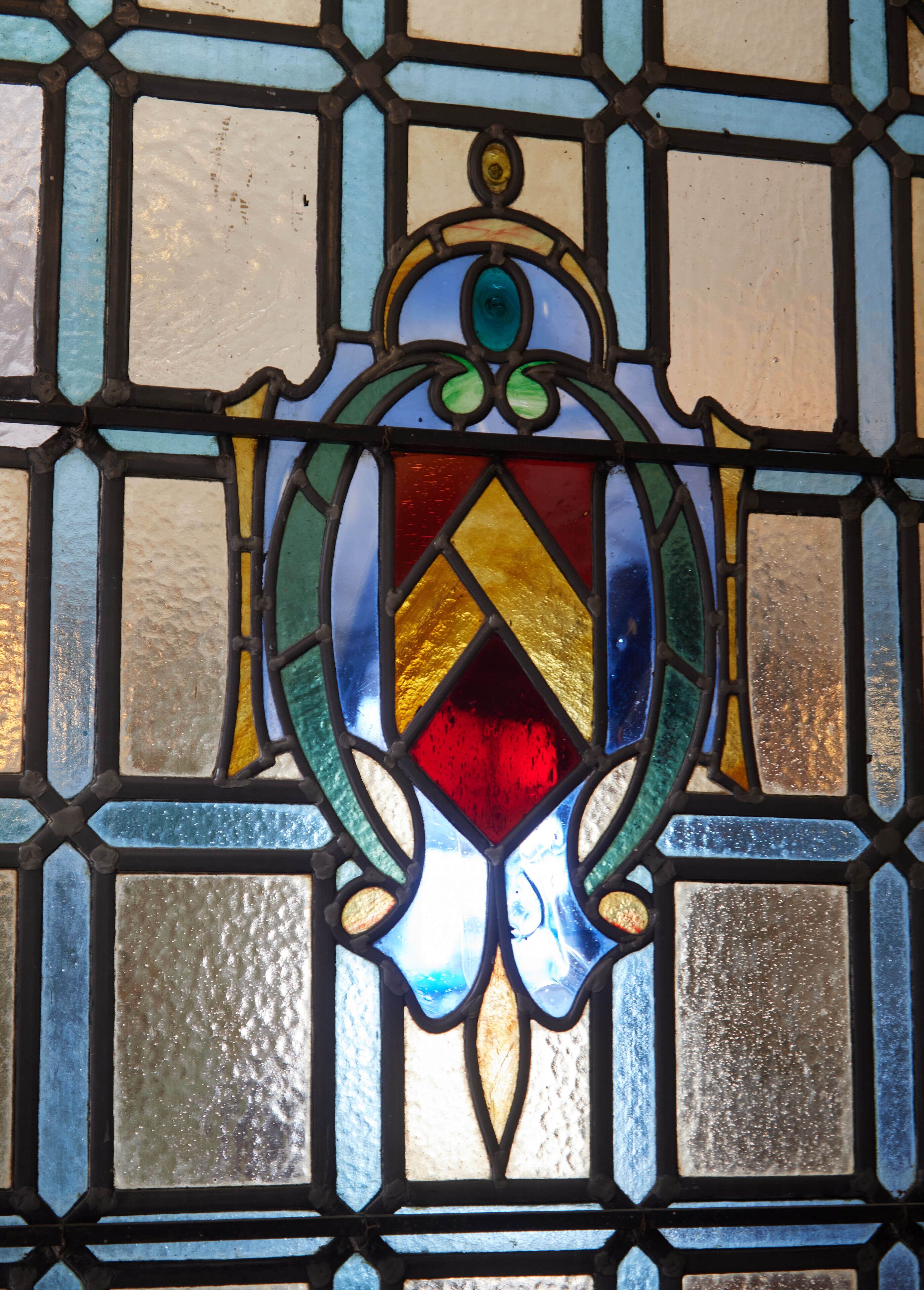 A stained glass window with a geometric design, featuring a central diamond shape with red and gold colors, surrounded by blue and green hues.
