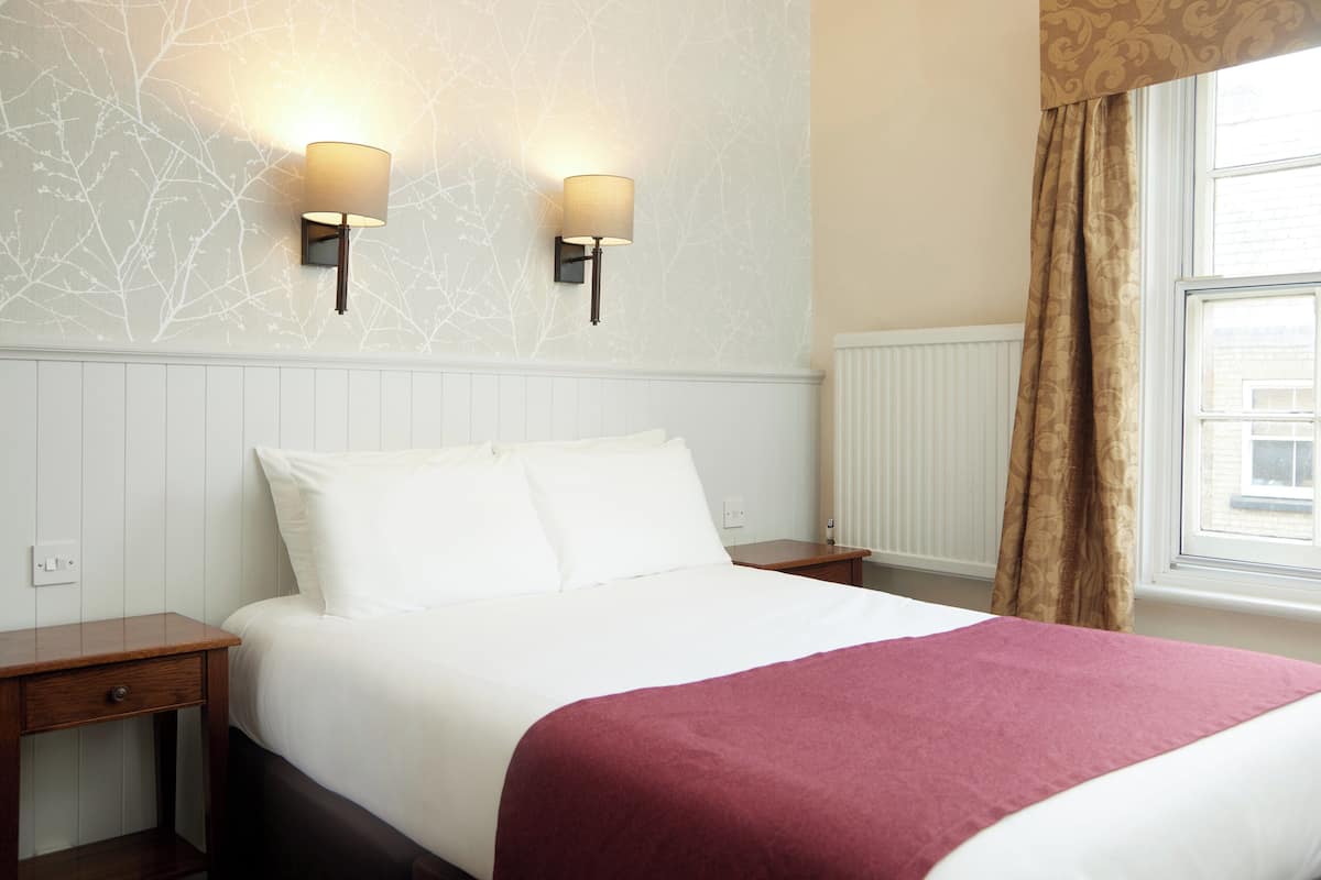A neatly made bed with white linens and a red throw blanket, two bedside tables with lamps, and a window with curtains.