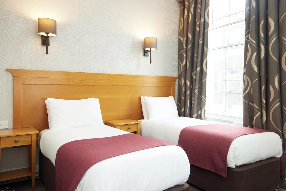 A hotel room with two beds, a wooden headboard, nightstands, wall-mounted lamps, patterned curtains, and a window with a view.