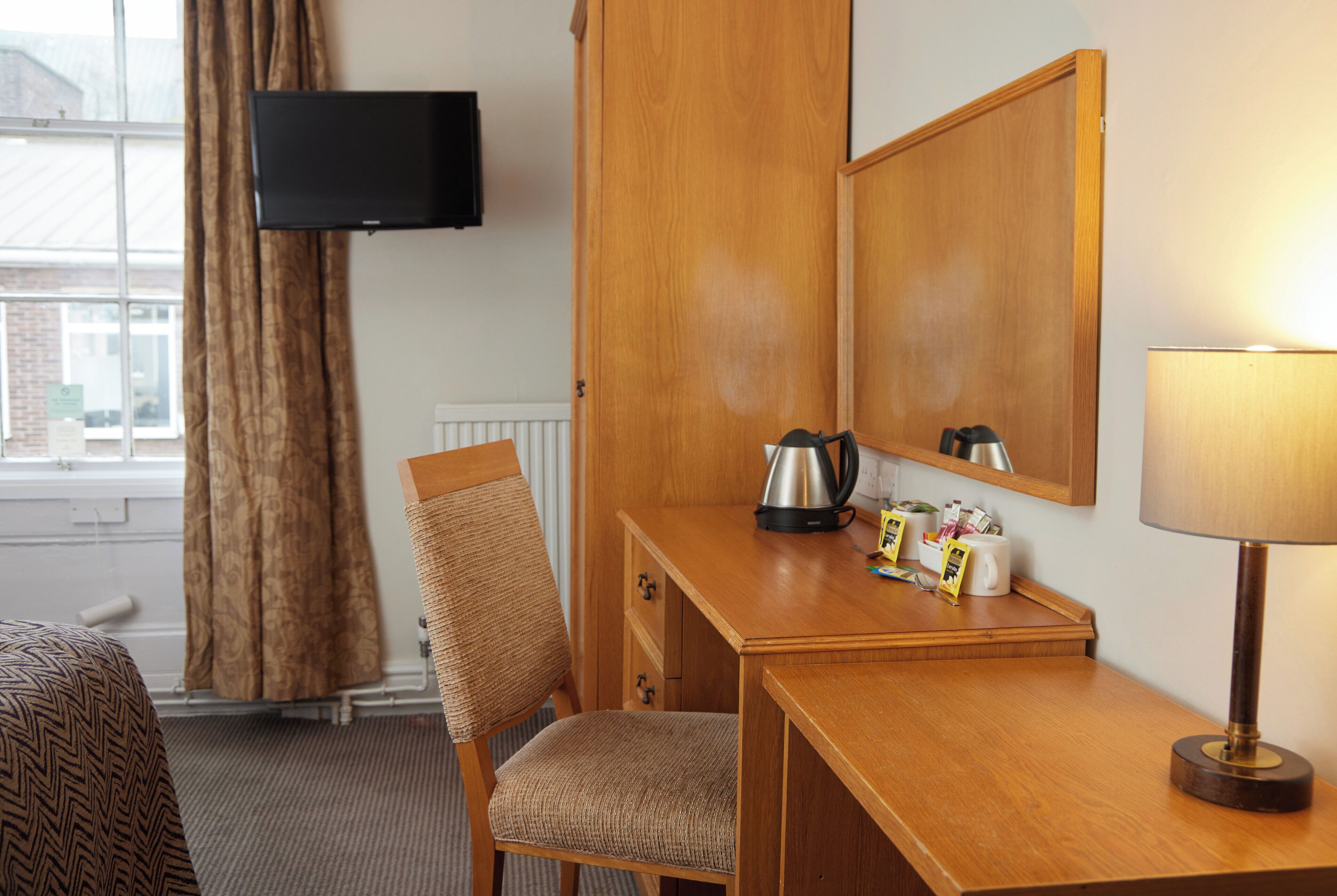 Une chambre d’hôtel avec un bureau en bois, une chaise, un téléviseur fixé au mur, une lampe et une fenêtre avec des rideaux.