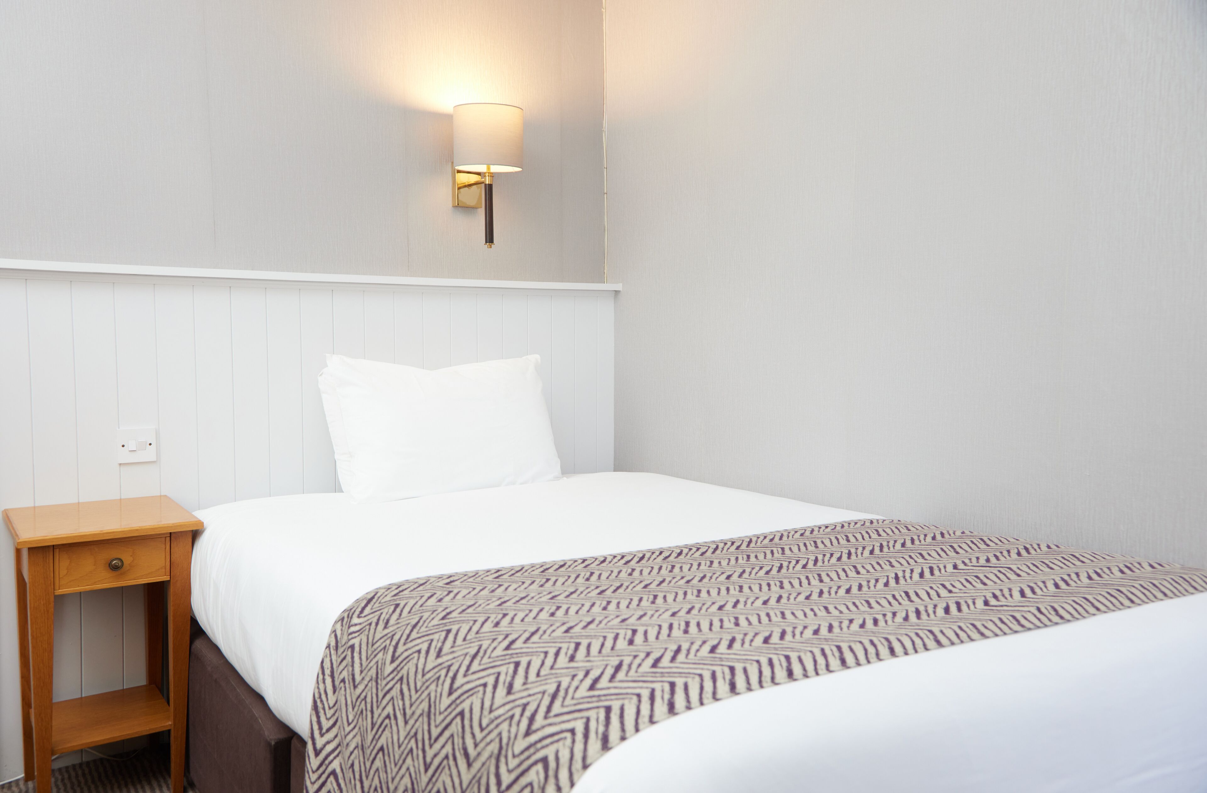 Habitación de hotel con una cama individual, una mesita de noche, una lámpara de pared y una manta decorativa.