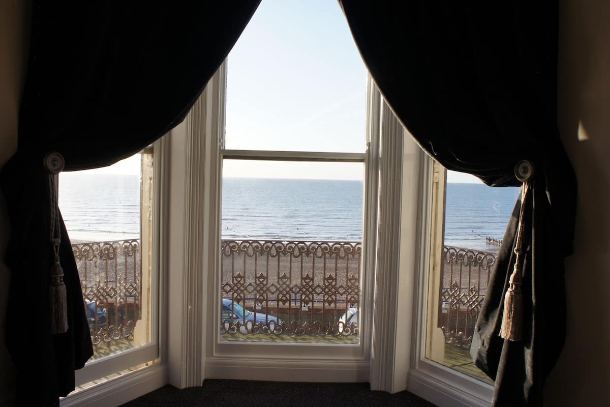 A corner room with a large window overlooking the sea, a balcony with a metal railing, and a door with a dark curtain.